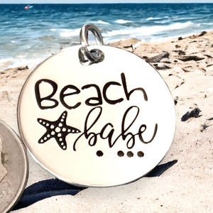 Puede incluir: Encanto de plata con una estrella de mar en blanco y negro y el texto "Beach babe" en escritura negra.