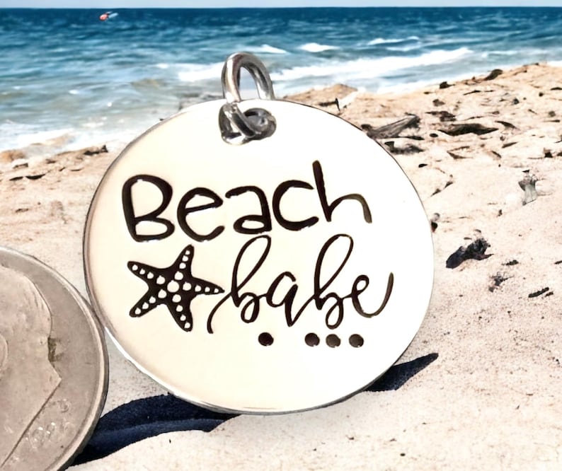 Puede incluir: Encanto de plata con una estrella de mar en blanco y negro y el texto "Beach babe" en escritura negra.