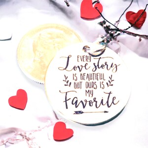 May include: White wooden circle with the text "Every love story is beautiful but ours is my favorite" with an arrow pointing to the right. The circle is surrounded by red hearts and white hearts on a white background.
