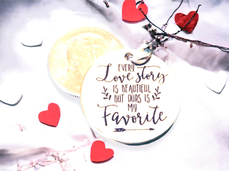May include: White wooden circle with the text "Every love story is beautiful but ours is my favorite" with an arrow pointing to the right. The circle is surrounded by red hearts and white hearts on a white background.