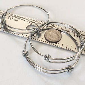 May include: Two silver bangle bracelets with knotted closures. The bracelets are laying on a ruler with a US quarter for scale.