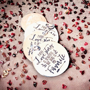 May include: Three round wooden tags with a white background and black calligraphy. The tags are strung together with a silver chain. The text on the tags reads "Every love story is of our favorite" and "Every love story is beautiful but ours is my favorite".
