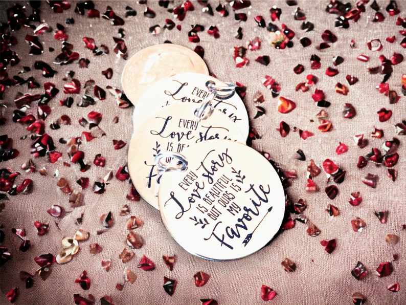 May include: Three round wooden tags with a white background and black calligraphy. The tags are strung together with a silver chain. The text on the tags reads "Every love story is of our favorite" and "Every love story is beautiful but ours is my favorite".