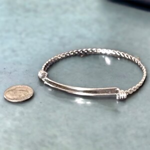 May include: A silver bracelet with a twisted rope design and a straight bar accent. The bracelet is next to a silver coin.