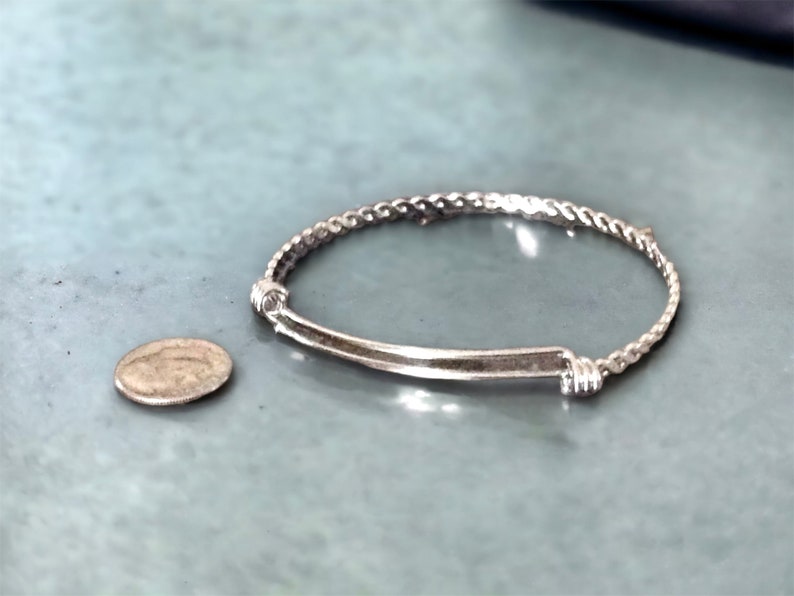 May include: A silver bracelet with a twisted rope design and a straight bar accent. The bracelet is next to a silver coin.