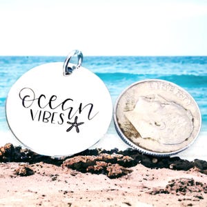 May include: A silver round charm with the words "Ocean Vibes" and a small starfish design. The charm is sitting on a sandy beach with a blurred background of the ocean.