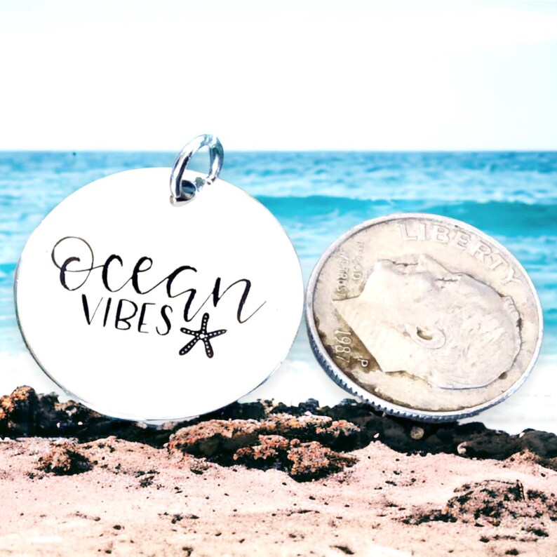 May include: A silver round charm with the words "Ocean Vibes" and a small starfish design. The charm is sitting on a sandy beach with a blurred background of the ocean.