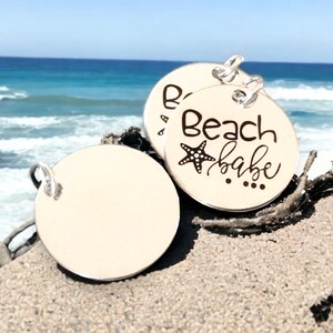 Puede incluir: Dos dijes redondos de plata con un anillo de fijaci&oacute;n. Un dije tiene el texto "Beach babe" con un dise&ntilde;o de estrella de mar. El otro dije est&aacute; en blanco.
