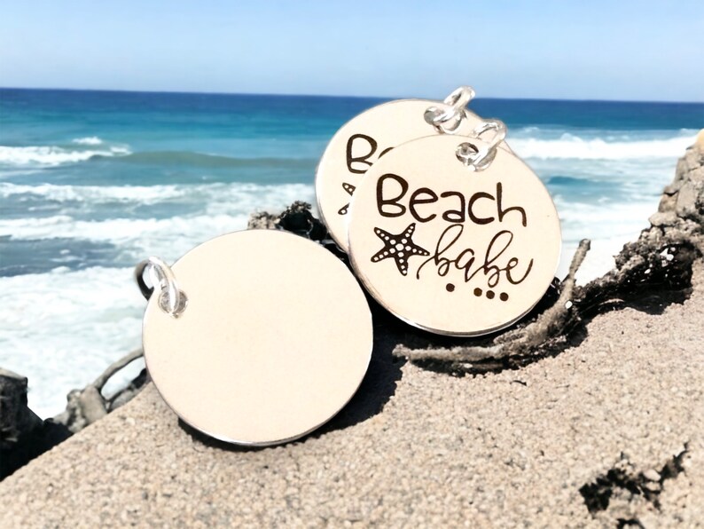 Puede incluir: Dos dijes redondos de plata con un anillo de fijaci&oacute;n. Un dije tiene el texto "Beach babe" con un dise&ntilde;o de estrella de mar. El otro dije est&aacute; en blanco.