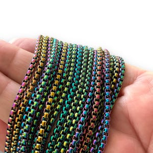 May include: A close-up of a multi-colored chain made of small metal links. The chain is iridescent, with shades of green, blue, purple, and gold. The chain is lying on a hand, and the light reflects off the metal links.