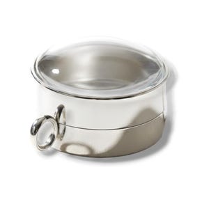 May include: A circular silver-toned metal container with a smooth, polished finish and a clear, domed glass lid. The container features a small, round metal ring attached to the side, serving as a handle or pull for the lid mechanism.