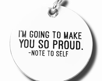 May include: A white, circular pendant with a silver-colored loop. The pendant features the black text: "I'M GOING TO MAKE YOU SO PROUD. -NOTE TO SELF."