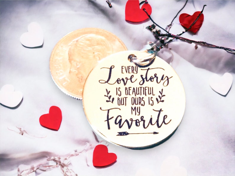 May include: A gold-colored round keychain with the text "Every love story is beautiful but ours is my favorite" engraved on it. The keychain has a small arrow design below the text.