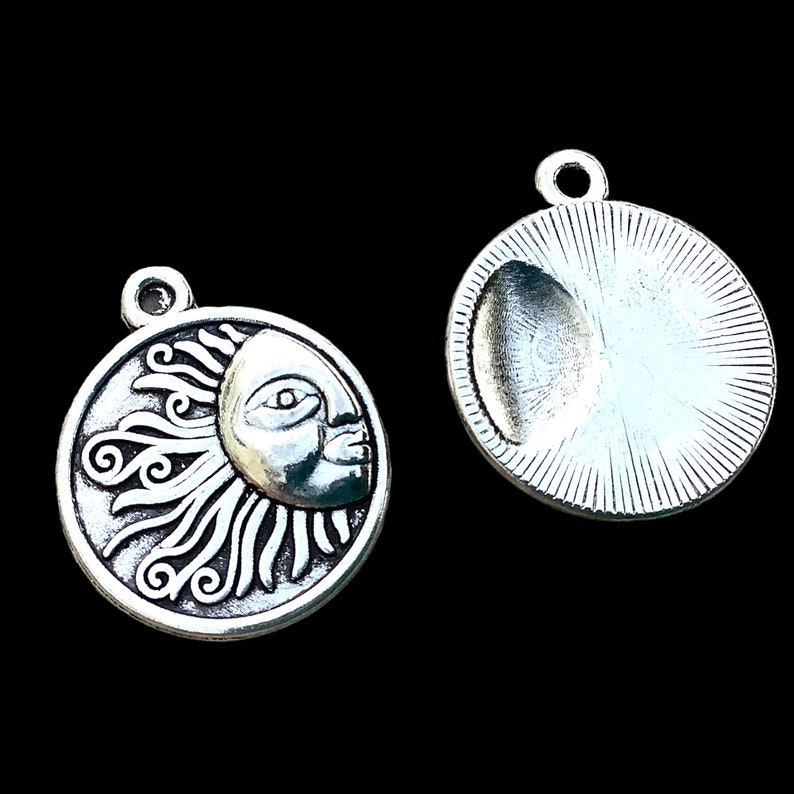 May include: Two silver-toned celestial charms. One charm features a sun with a face and swirling rays. The other charm has a blank, concave surface with a sunburst design on the back. Both charms have a loop for attaching to jewelry.