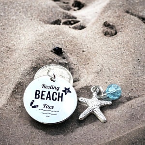 May include: Silver charm with the text "Resting Beach Face" and a small starfish. The charm is on a sandy surface with a blue glass bead.