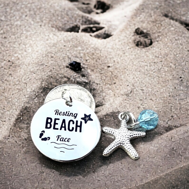 May include: Silver charm with the text "Resting Beach Face" and a small starfish. The charm is on a sandy surface with a blue glass bead.