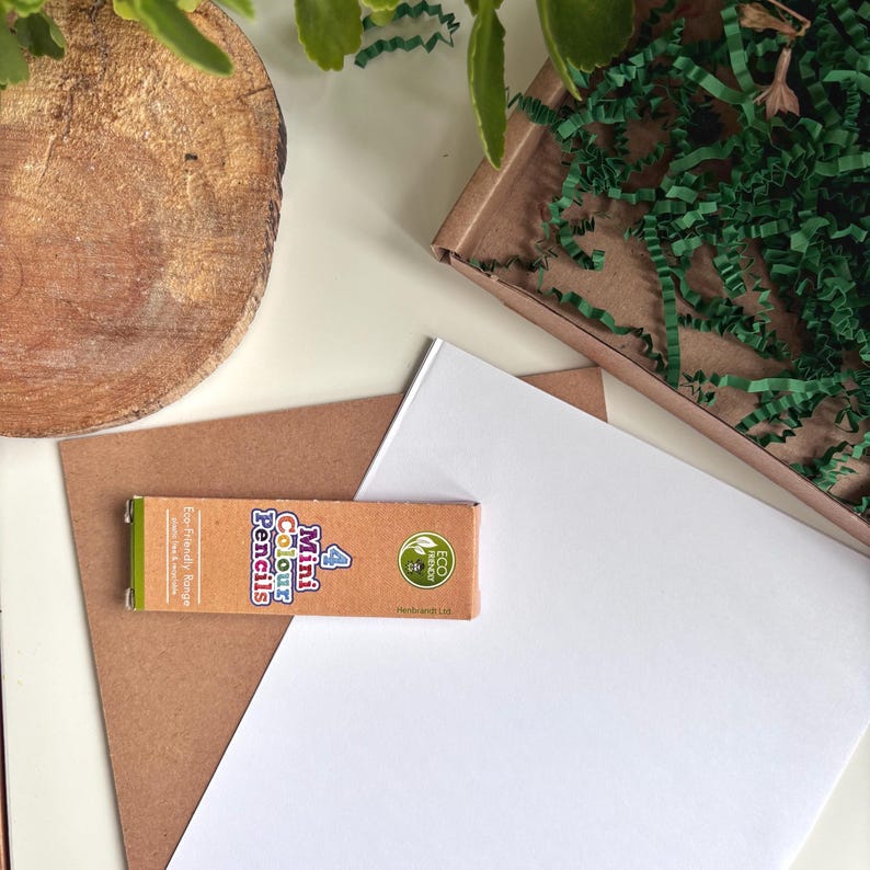 A flat lay arrangement showing a white sheet of paper, a brown card, and a small box of 4 Mini Colour Pencils. The scene is decorated with green leaves and a wooden disc arranged on a white background.