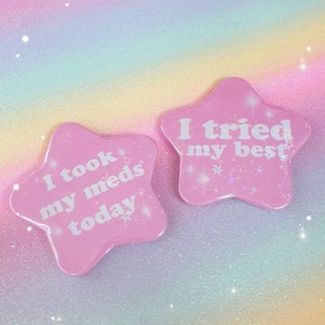 Può includere: Due spille rosa a forma di stella con testo bianco. Una dice "I took my meds today", l'altra "I tried my best". Le spille hanno una finitura lucida e sono decorate con piccole stelle e glitter. Lo sfondo è un gradiente arcobaleno pastello.