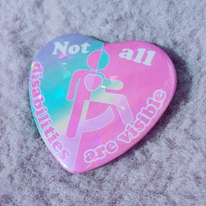 May include: Heart-shaped button with a split design. One half is light blue and the other is pink. The button features the text "Not all disabilities are visible" and a graphic of a person in a wheelchair.