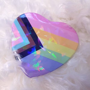 May include: A heart-shaped compact with a rainbow design. The design features a holographic finish with a variety of colors, including pink, blue, purple, and green.