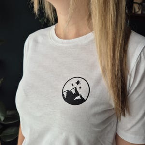 Könnte beinhalten: Weißes T-Shirt mit einem schwarzen, kreisförmigen Grafikdesign, das eine Bergkette und Sterne zeigt. Das Design befindet sich auf der Brust. Das T-Shirt ist aus weichem Material.
