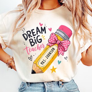 Puede incluir: Una camiseta de color crema con un gráfico de un lápiz amarillo con un lazo rosa de lunares, el texto "DREAM BIG teacher" en negro y rosa, y el nombre "MRS. JOHNSON" impreso en el lápiz, rodeado de pequeños corazones y una estrella.