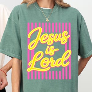 Puede incluir: Una camiseta de manga corta color verde salvia con un diseño gráfico de rayas verticales rosa intenso y el texto amarillo "Jesus is Lord" en una fuente cursiva. La prenda tiene cuello redondo y una silueta holgada y relajada.