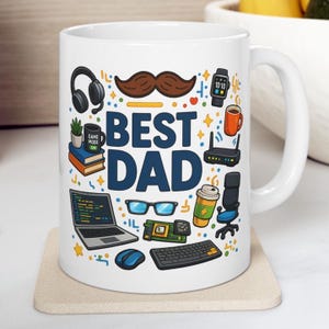 Puede incluir: Taza de cerámica blanca con la frase "BEST DAD" en azul, rodeada de ilustraciones de un bigote, auriculares, portátil, gafas y otros elementos tecnológicos. La taza está sobre un posavasos beige.