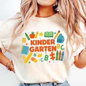 May include: Cream-colored t-shirt with a colorful "Kindergarten" graphic. The design includes a red apple, crayons, a backpack, and a book. The shirt is a casual style, suitable for back-to-school or everyday wear.