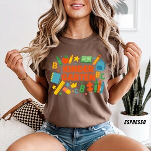 May include: Espresso-colored t-shirt with the words "KINDER GARTEN" in orange and green, surrounded by colorful school-themed graphics. The design includes crayons, a backpack, and a book.
