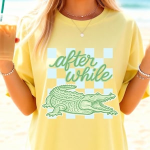 Puede incluir: Camiseta amarilla con un gráfico de cocodrilo verde y la frase "after while". El diseño incluye un patrón de cuadros en azul claro y blanco. La camiseta es de estilo casual.