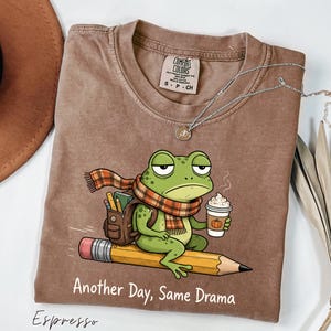 Puede incluir: Una camiseta marrón de manga corta con el gráfico de una rana verde con bufanda de cuadros y mochila, montada sobre un lápiz amarillo mientras sostiene un café. El texto "Another Day, Same Drama" aparece impreso en color blanco debajo.
