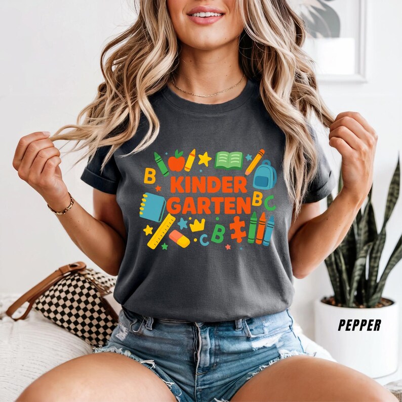 May include: Dark gray t-shirt with the words "KINDERGARTEN" in orange and green, surrounded by colorful school-themed graphics. The shirt features a crew neck and short sleeves. The model is wearing blue denim shorts.