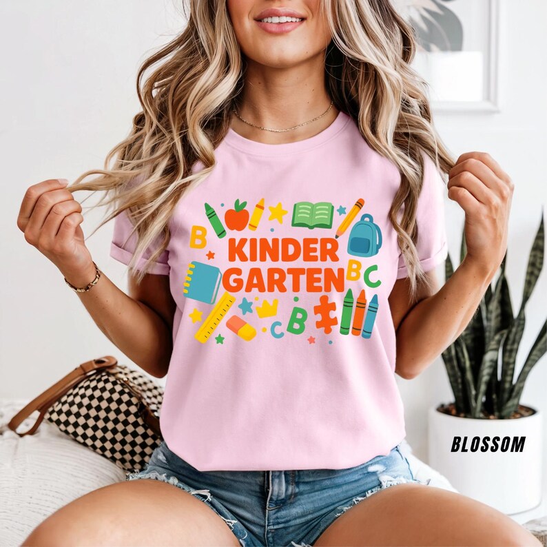 May include: A light pink t-shirt with the words "KINDERGARTEN" in orange and green, surrounded by colorful school supplies. The design includes a pencil, apple, crayons, and a backpack. The shirt is short-sleeved.