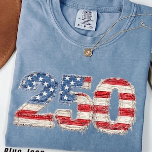 Puede incluir: Camiseta azul vaquero con el número "250" aplicado en la parte delantera con un diseño de bandera americana desgastada. Un collar dorado con un colgante redondo descansa sobre la camiseta. La camiseta tiene una etiqueta Comfort Colors.