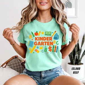 May include: A light green t-shirt with the words "KINDER GARDEN" in orange and blue, surrounded by school-themed graphics. The design includes a backpack, crayons, and an apple. The shirt is a short-sleeved crew neck.