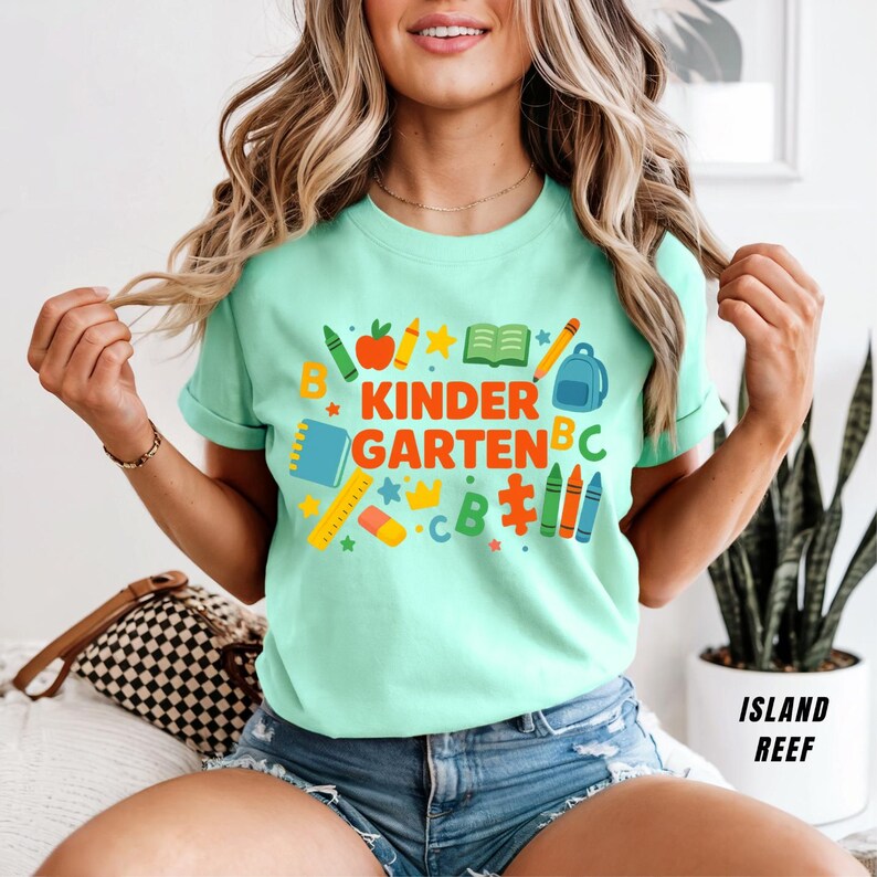 May include: A light green t-shirt with the words "KINDER GARDEN" in orange and blue, surrounded by school-themed graphics. The design includes a backpack, crayons, and an apple. The shirt is a short-sleeved crew neck.