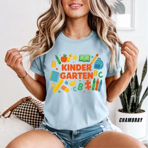 May include: Light blue t-shirt with the words "KINDER GARDEN" in orange and green, surrounded by colorful school-themed graphics. The design includes crayons, an apple, a book, a backpack, and a ruler. The shirt is made of a soft material.