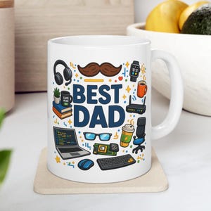 Puede incluir: Taza de cerámica blanca con un diseño colorido que dice "BEST DAD". La taza presenta ilustraciones de aparatos tecnológicos, un bigote y una taza de café. La taza está sobre un posavasos beige.