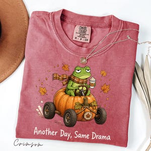May include: A crimson crewneck t-shirt featuring a graphic of a green frog wearing a plaid scarf, driving a pumpkin car while holding a coffee cup. The design includes falling autumn leaves and the text Another Day, Same Drama printed below.