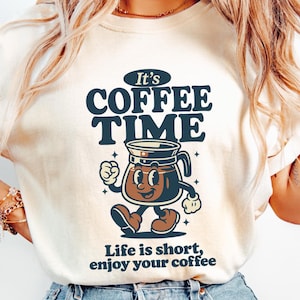May include: A cream-coloured sweatshirt with the navy blue text "It's COFFEE TIME". Below the text is a cartoon coffee pot with a smiling face and legs. The phrase "Life is short, enjoy your coffee" is printed at the bottom of the design.