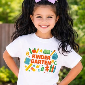 May include: A young girl with long black hair and a light purple bow smiles, wearing a white t-shirt. The shirt features the words "Kindergarten" in colorful letters, along with school-themed graphics like crayons, an apple, and a backpack.