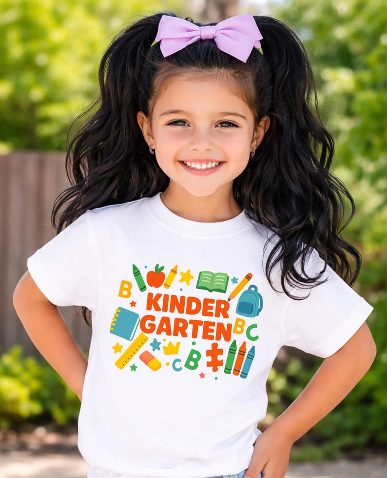 May include: A young girl with long black hair and a light purple bow smiles, wearing a white t-shirt. The shirt features the words "Kindergarten" in colorful letters, along with school-themed graphics like crayons, an apple, and a backpack.