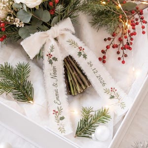 May include: A white gift box containing a bouquet of white roses, red berries, and evergreen sprigs. The bouquet is tied with a cream-colored ribbon featuring the printed text "Emma & Noah" and "24.12.2026" along with small holly illustrations.