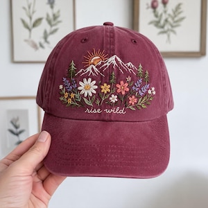 Op de afbeelding: Een bordeauxrode baseballpet met een borduursel van een berglandschap met een zon, dennenbomen en kleurrijke wilde bloemen. De tekst "rise wild" is in witte cursieve letters onder het bloemmotief op het voorpaneel geborduurd.