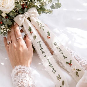May include: A floral bouquet featuring white roses, red berries, and evergreen sprigs, accented with two cream-colored fabric ribbons. The ribbons are embroidered with green holly leaves, red berries, and the text Guillermo & Marialy 26.09.2026.