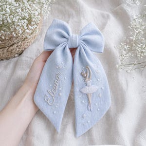 May include: A pale blue hair bow with embroidered details. One side features the name "Eleanor" in cursive, while the other side has a ballerina design. The bow is tied in the centre and has long, flowing tails.