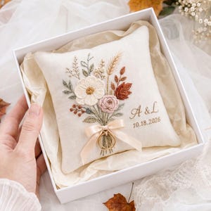 May include: A square off-white ring bearer pillow featuring an embroidered floral bouquet in shades of cream, pink, and rust. The pillow includes a cream satin ribbon for securing rings and is personalized with the initials A & L and the date 10.18.2026.