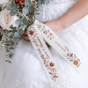 Puede incluir: Un ramo de novia de rosas blancas, astilbe rojo y hojas de eucalipto atado con dos cintas color crema deshilachadas. Las cintas presentan bordados de follaje otoñal, setas y calabazas, con el texto Laura & Alexandre y la fecha 26.09.2026.