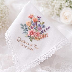 Op de afbeelding: Een witte zakdoek met een kanten rand voorzien van een geborduurd bloemenboeket in roze, oranje, blauwe en groene tinten. Onder de bloemen staat de tekst Quitterie et Tom en de datum 19.09.2026 in bruin cursief borduurwerk.